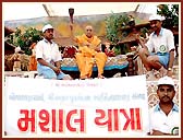 Swamishri holding aloft a torch carried by a team of 31 youths who pilgrimaged on foot from Amdavad to Sarangpur (156 Km)