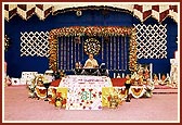 Swamishri engaged in his morning puja with a backdrop of flower decorations and a giant cake in the foreground