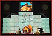 Swamishri seated with a colourful backdrop on stage  