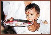 A child first curiously looks on and then joins his father in performing the patotsav arti