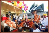 Swamishri, sadhus and devotees engaged in devotional singing, with the Sydney Opera House in the background