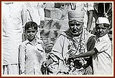 Swamishri garlanded by a balak Swanand Pandurang Patel, and senior devotees - Shri Kaniayalal Kishan, Shri Sukhlal Vithal (black topee) and Shri Shaligram Ramdas (far left) outside the Swaminarayan Mandir in Kusumbe, January 1976